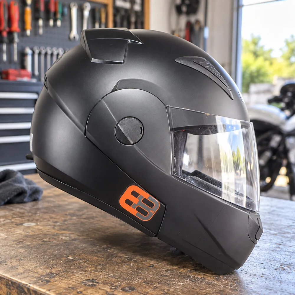 Capacete articulado preto fosco com logotipo laranja na lateral, sobre uma bancada de madeira com ferramentas ao fundo