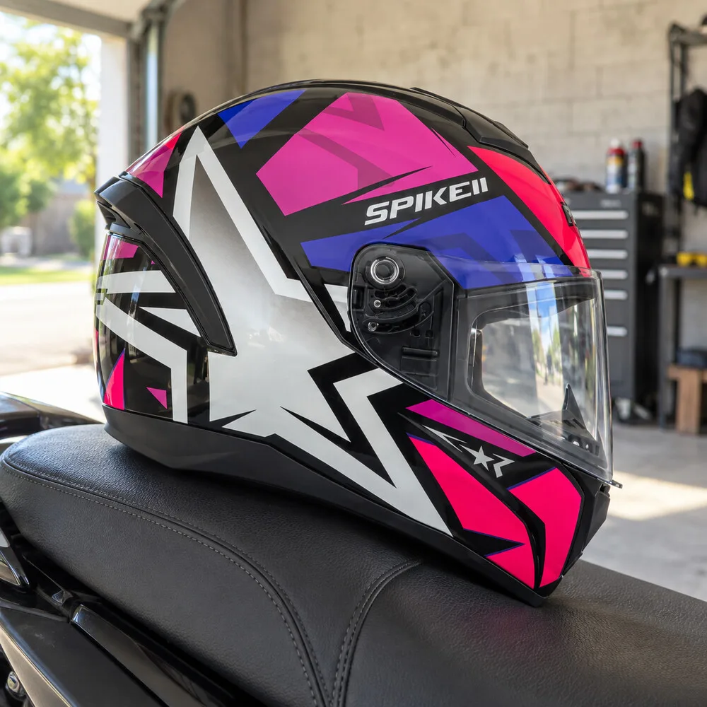 Capacete fechado com grafismo de estrela em rosa, branco e roxo, apoiado no banco de uma moto numa garagem